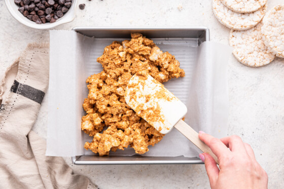 A woman's hand uses a silicone spatula to spread the ingredients for the no bake peanut butter crunch bars in a square baking dish.