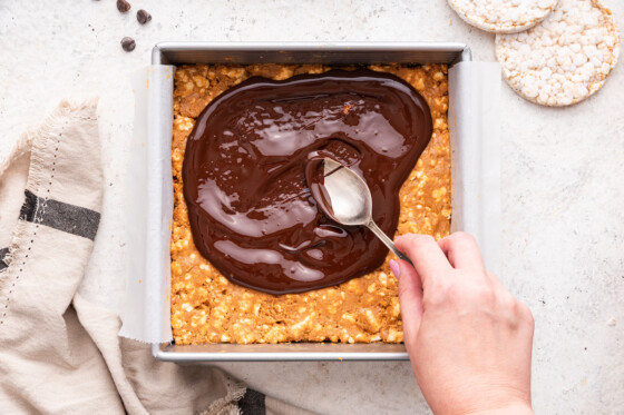 A woman's hand using a spoon to spread melted chocolate over the no bake peanut butter crunch bars.