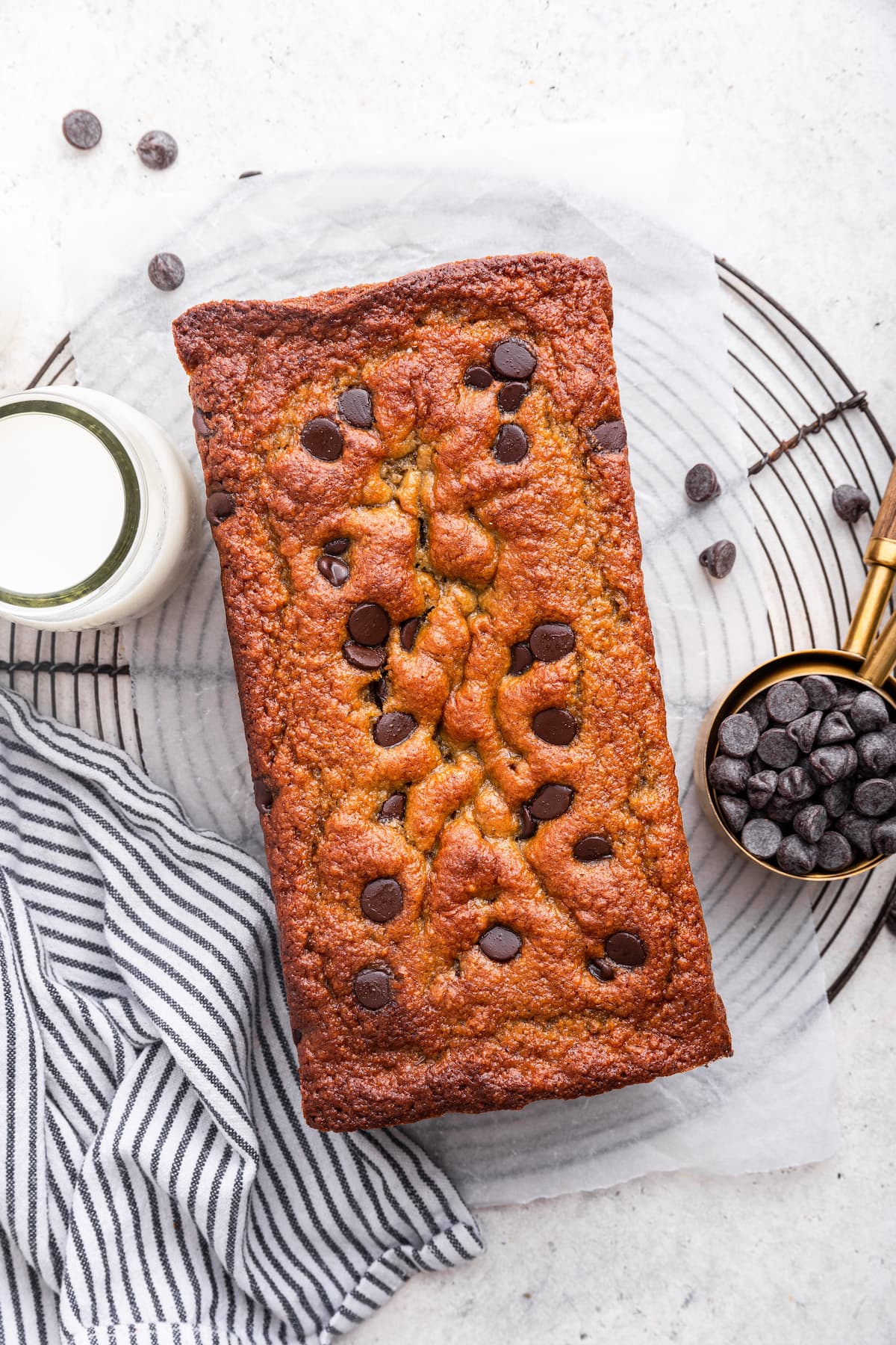 A loaf of protein banana bread with chocolate chips near a glass of milk and a small measuring cup of chocolate chips.