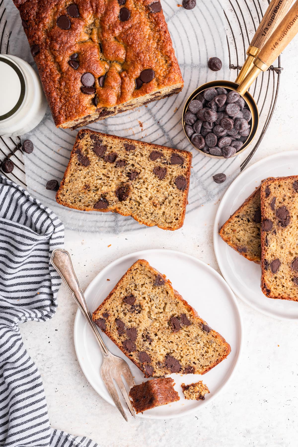 A loaf of protein banana bread with chocolate chips near a glass of milk and a small measuring cup of chocolate chips. There's two small plates with sliced banana bread on them with one of them having a fork taking a small bit size piece.