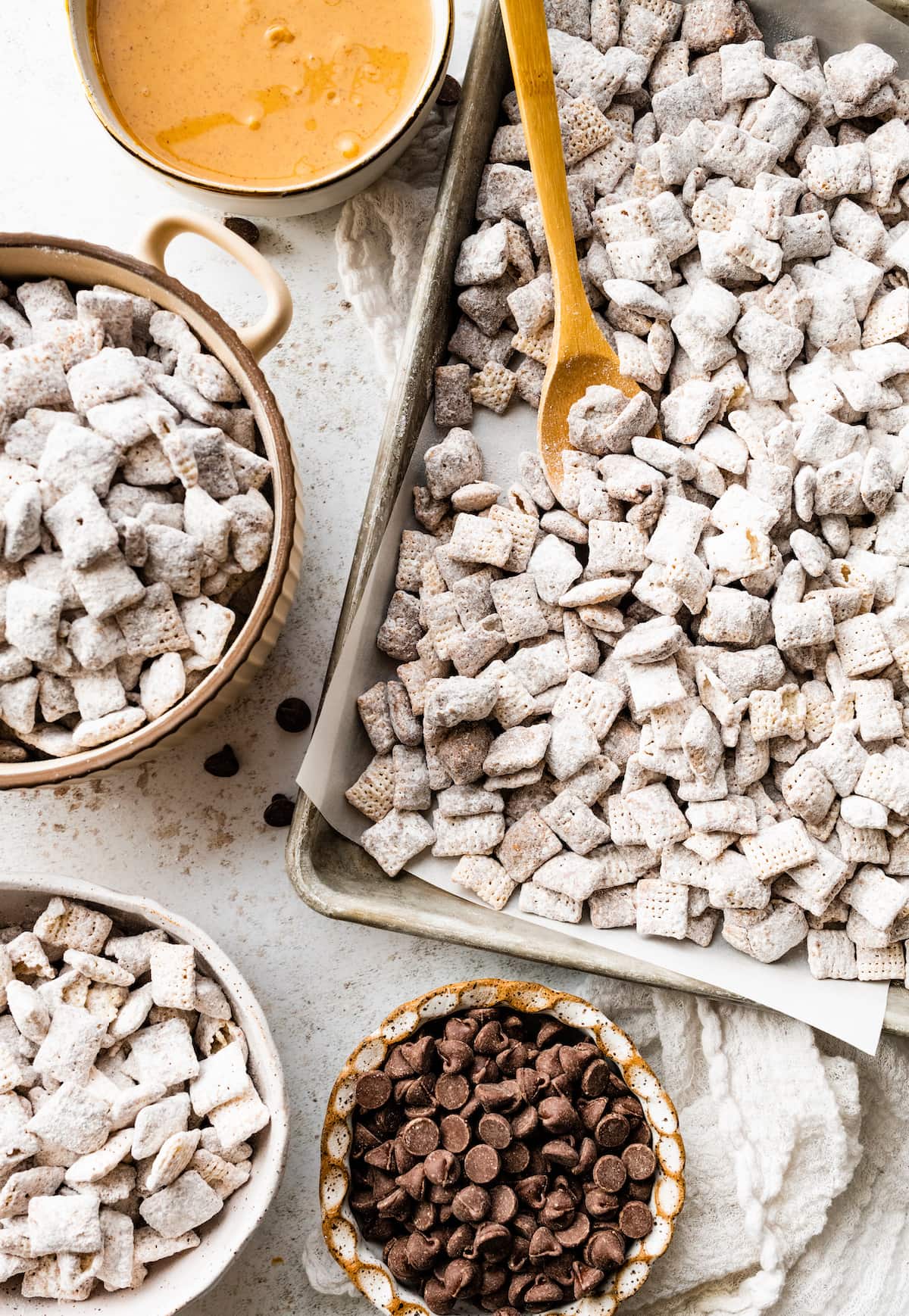 Puppy chow on a baking tray with a wooden spoon near one small bowl of chocolate chips, another small bowl of peanut butter, and other small bowls with the puppy chow.