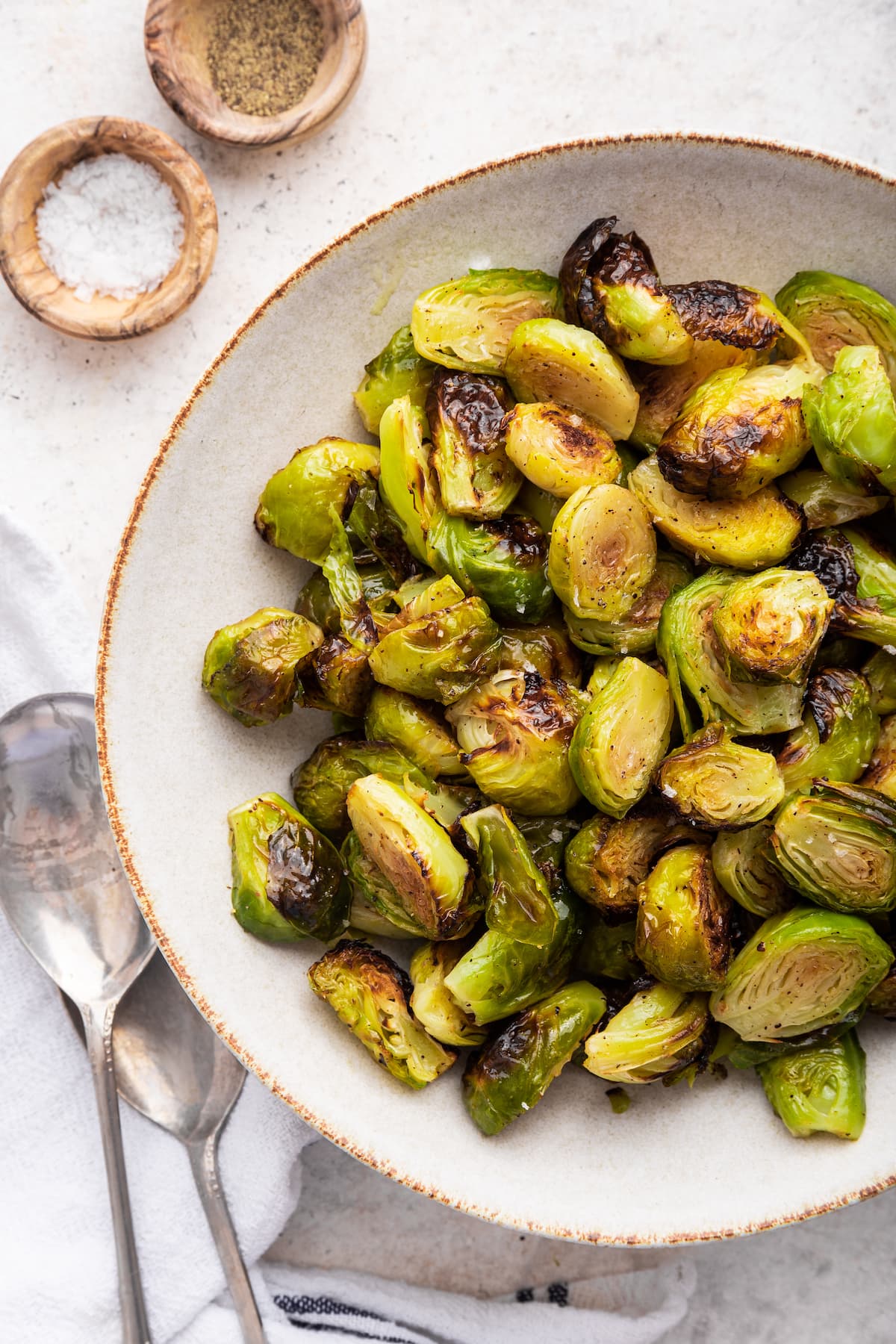 Roasted brussles sprouts in a large bowl near two serving spoons and two small wooden bowls containing salt and pepper.