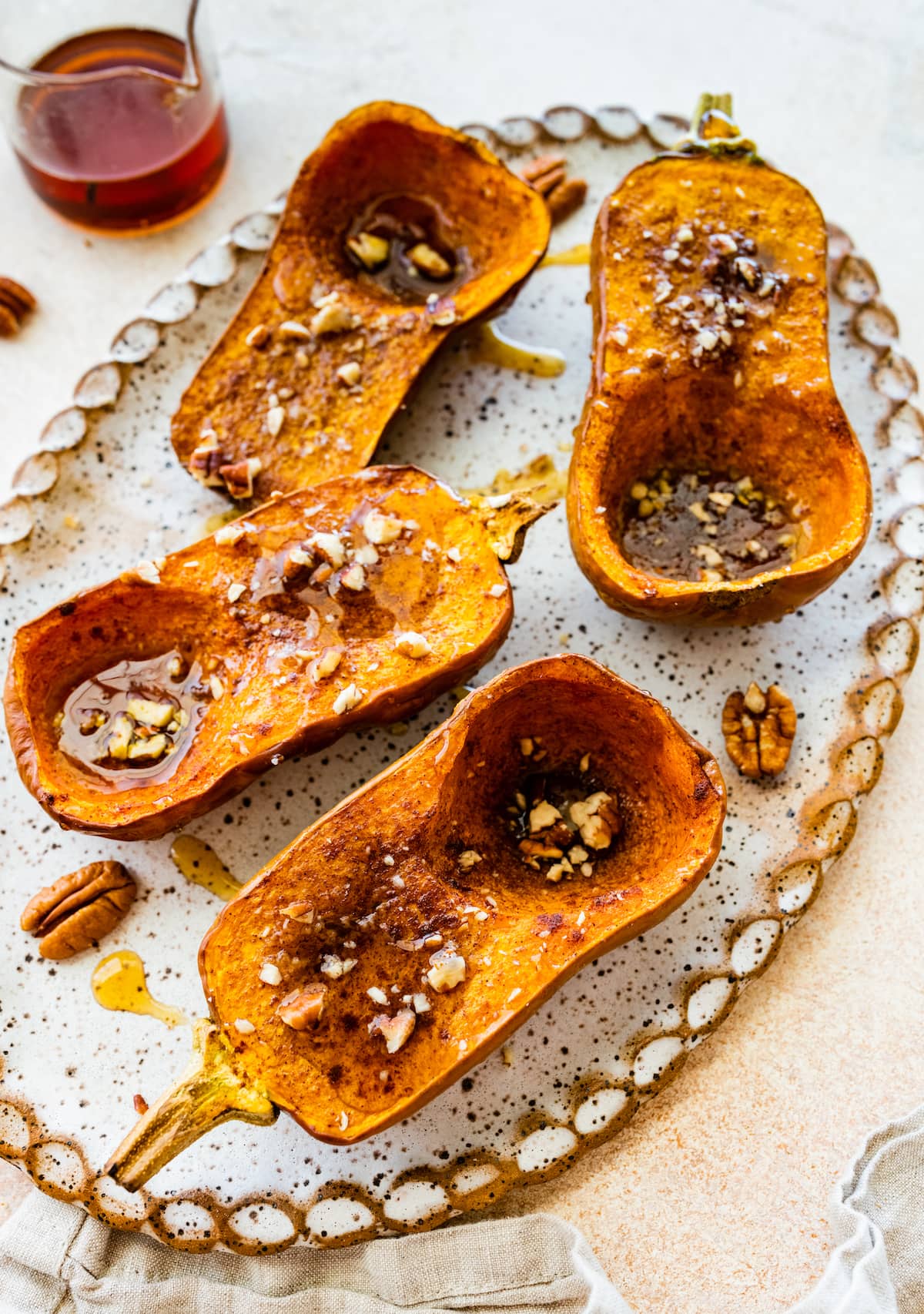 Two halved roasted honeynut squash on a large serving plate with maple syrup and crushed pecans.