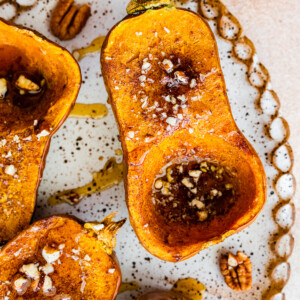 Half of a roasted honeynut squash on a large serving plate topped with maple syrup, crushed pecan, and sea salt.