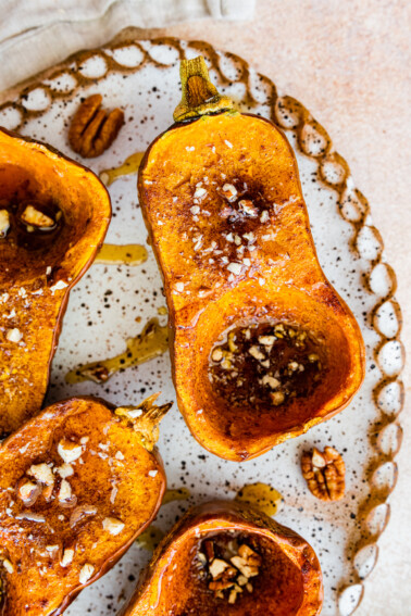 Half of a roasted honeynut squash on a large serving plate topped with maple syrup, crushed pecan, and sea salt.