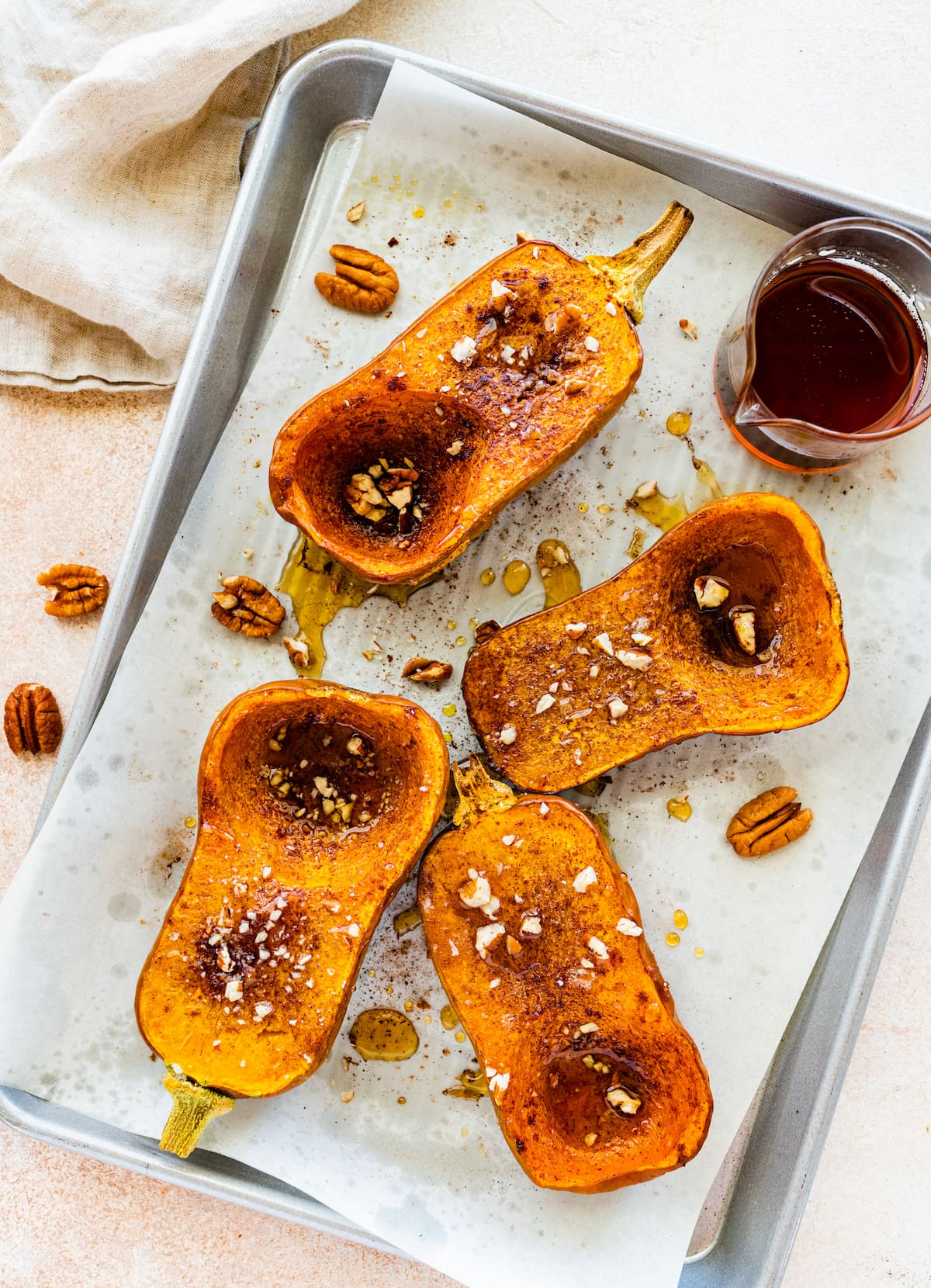 Two halved roasted honeynut squash on a baking tray with maple syrup and crushed pecans.