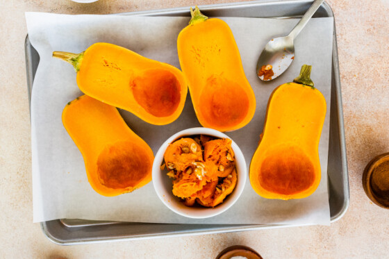 Two honeynut squashes cut in half and deseeded on a baking tray near a large metal spoon and a small bowl containing the seeds of the squash.