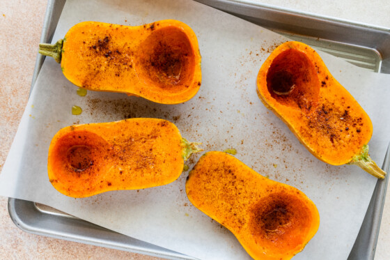 Two honeynut squashes cut in half on a baking tray covered in maple syrup and cinnamon before being roasted.