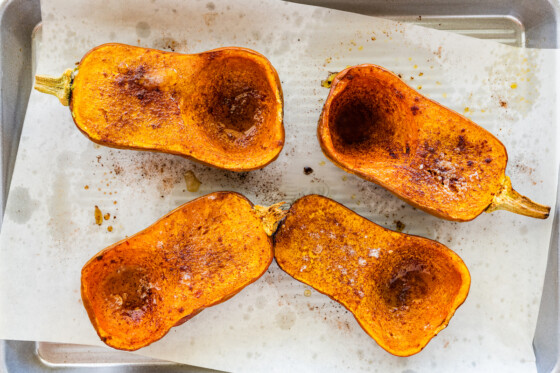 Two honeynut squashes cut in half on a baking tray covered in maple syrup and cinnamon after being roasted.