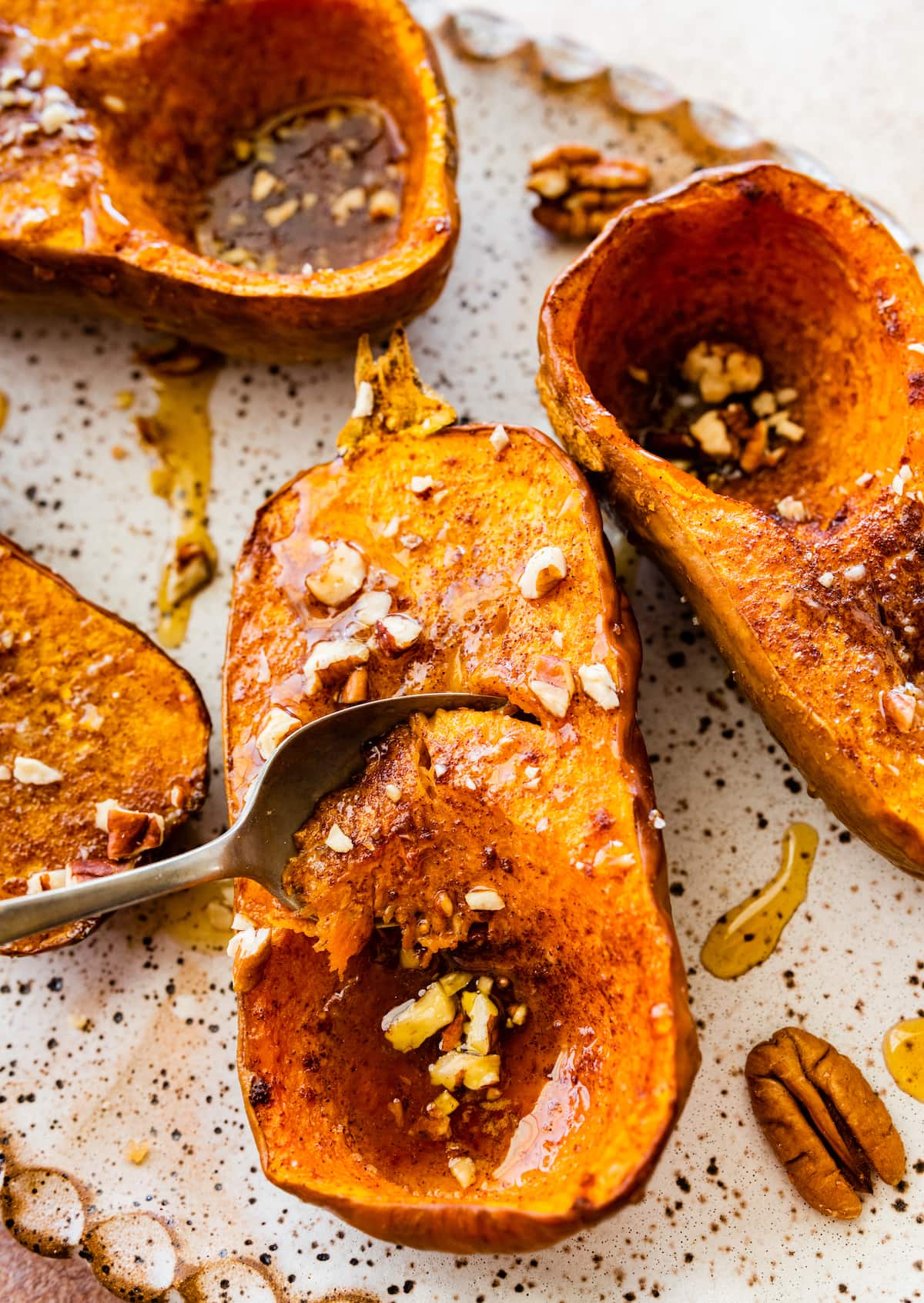 A large spoon taking a scoop of roasted honeynut squash that is on a large plate and covered in maple syrup, crushed pecans, cinnamon, and salt.