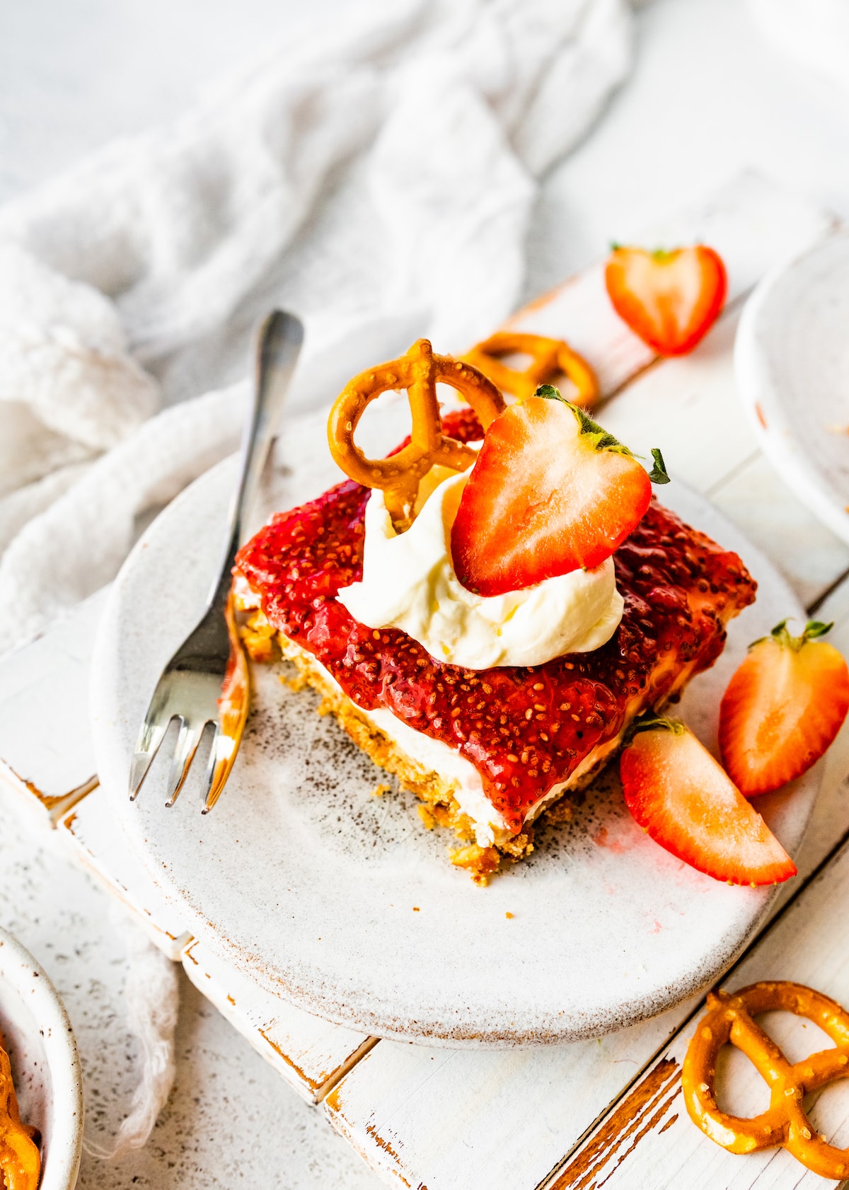 A slice of strawberry pretzel salad on a small plate with a metal fork. The slice is topped with a dollop of whipped cream, a sliced strawberry, and a pretzel.