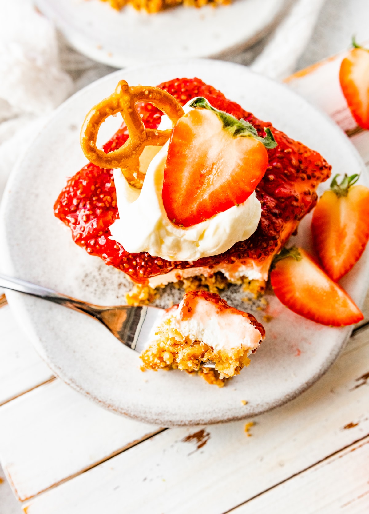 A slice of strawberry pretzel salad on a small plate with a metal fork holding a bite-size piece. The slice is topped with a dollop of whipped cream, a sliced strawberry, and a pretzel.