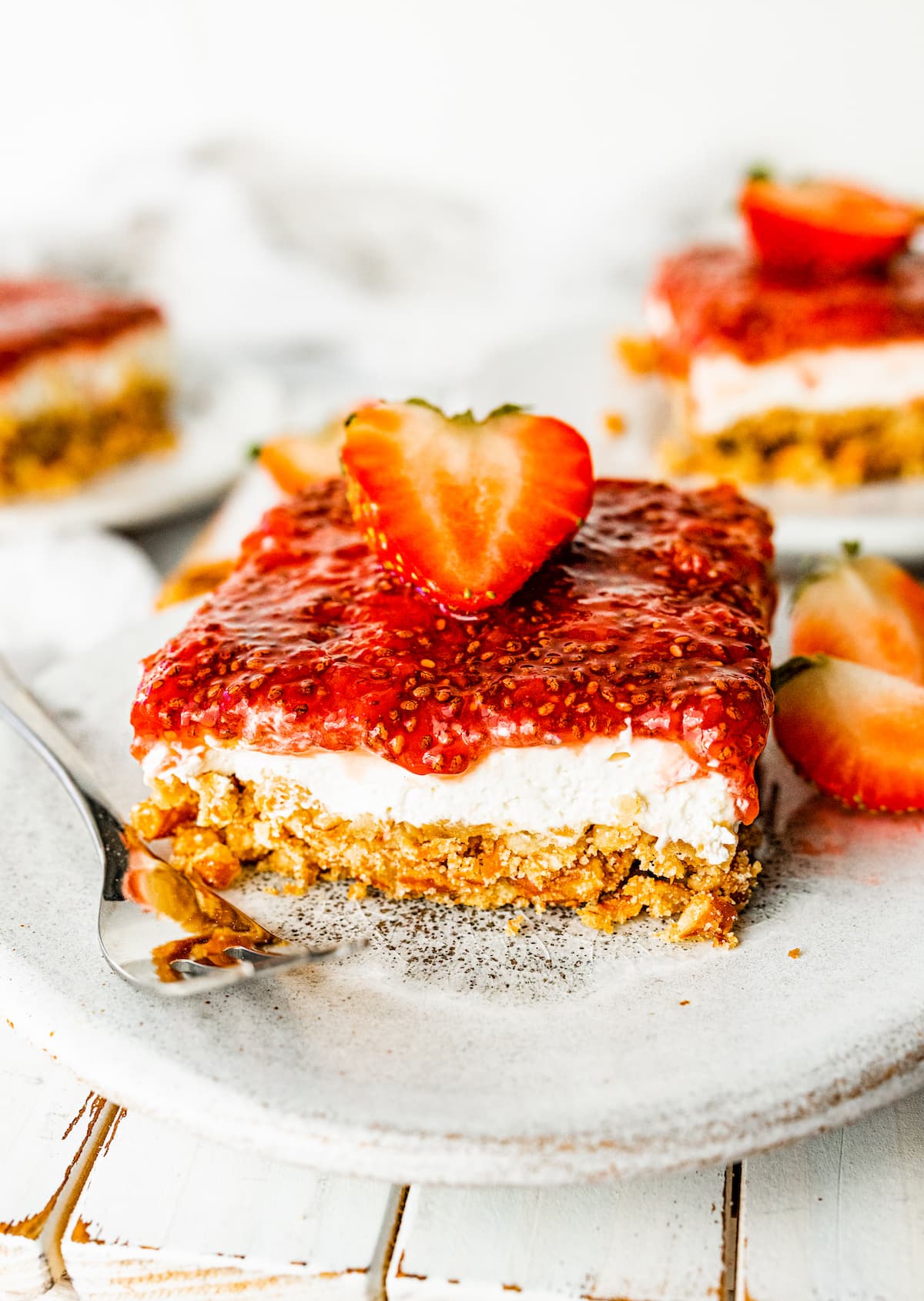 A slice of strawberry pretzel salad on a small plate with a metal fork. The slice is topped with a sliced strawberry.