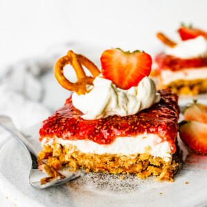 A slice of strawberry pretzel salad on a small plate with a metal fork. The slice is topped with a dollop of whipped cream, a sliced strawberry, and a pretzel.