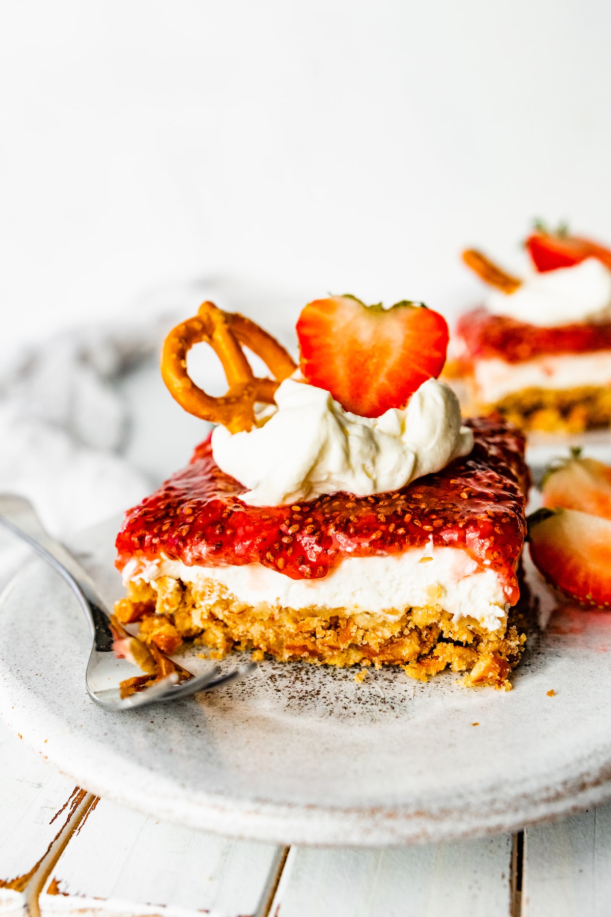 A slice of strawberry pretzel salad on a small plate with a metal fork. The slice is topped with a dollop of whipped cream, a sliced strawberry, and a pretzel.