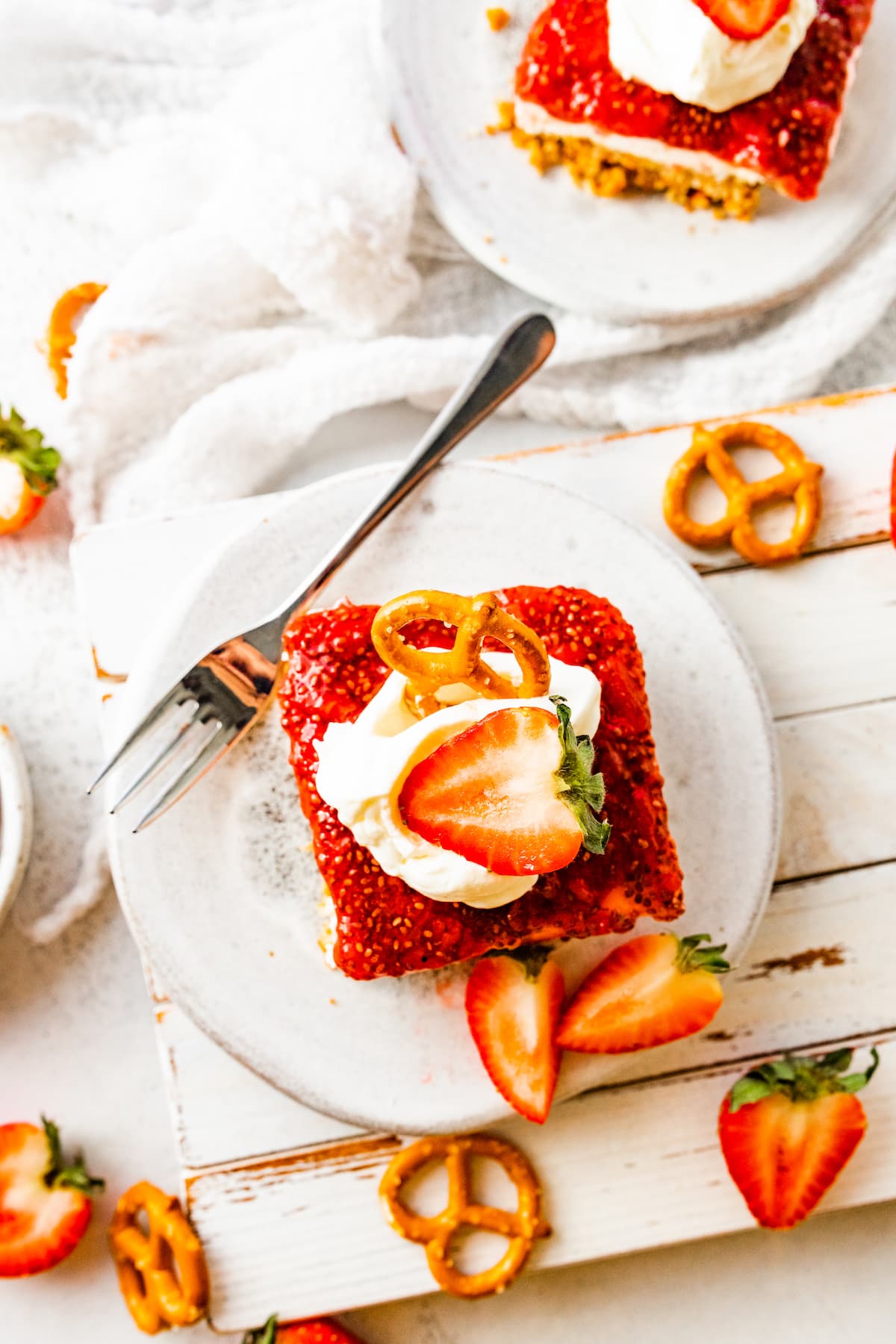 A slice of strawberry pretzel salad on a small plate with a metal fork. The slice is topped with a dollop of whipped cream, a sliced strawberry, and a pretzel.