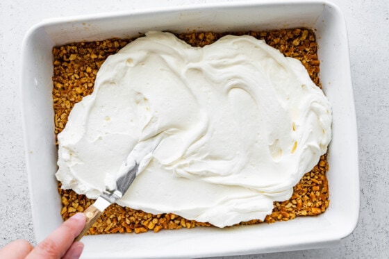 A woman's hand spreading the whipped cream cheese and yogurt mixture over a bed of crushed pretzels in a large baking dish.