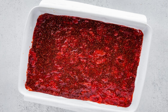 A layer of strawberry chia jam spread out in a large baking pan over a layer of a cream cheese and yogurt mixture and a bottom layer of crushed pretzels in a large baking dish.