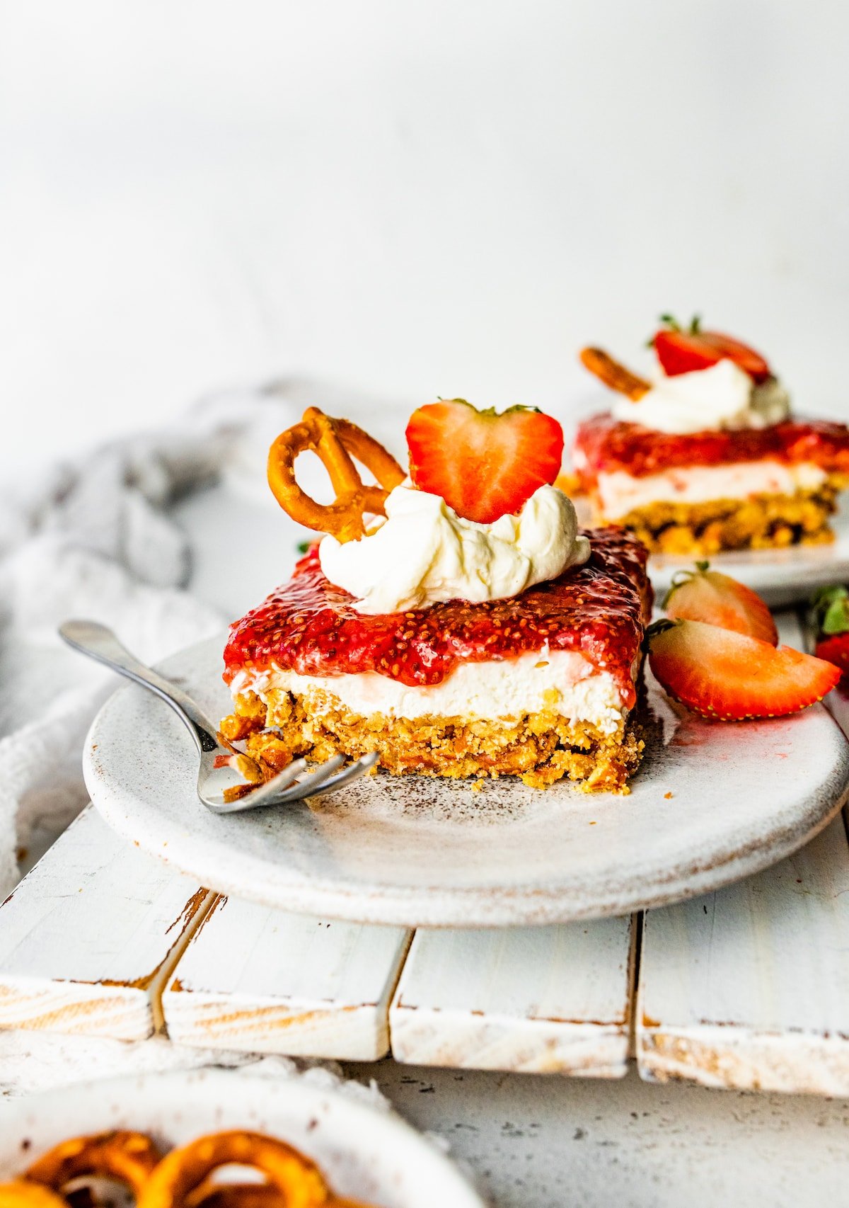 A slice of strawberry pretzel salad on a small plate with a metal fork. The slice is topped with a dollop of whipped cream, a sliced strawberry, and a pretzel.