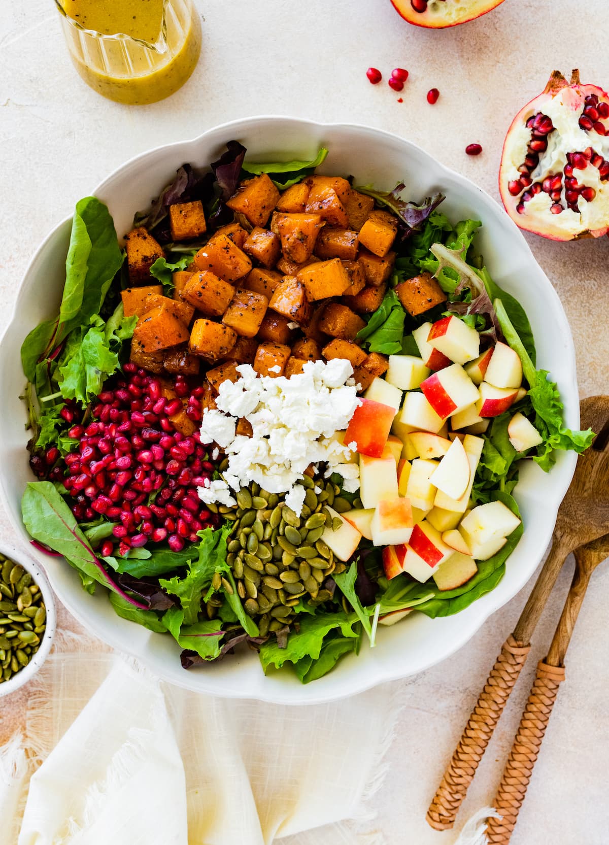 Thanksgiving salad in a large bowl with the ingredients that include butternut squash, apple, goat cheese, pomegranate, pumpkin seeds, and greens.