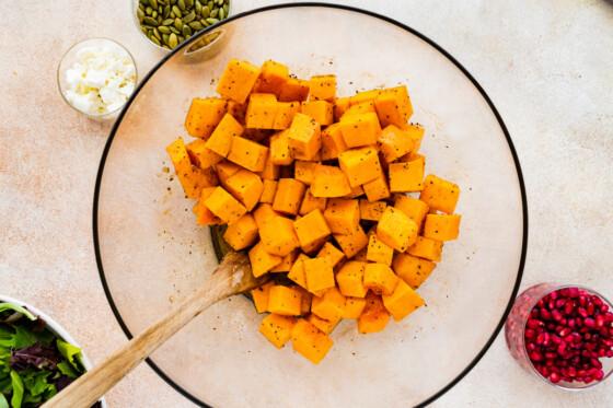 Cubed butternut squash in a large glass bowl with black pepper and a wooden spoon.