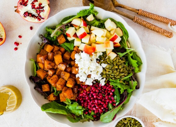 Thanksgiving salad in a large bowl with the ingredients that include butternut squash, apple, goat cheese, pomegranate, pumpkin seeds, and greens.