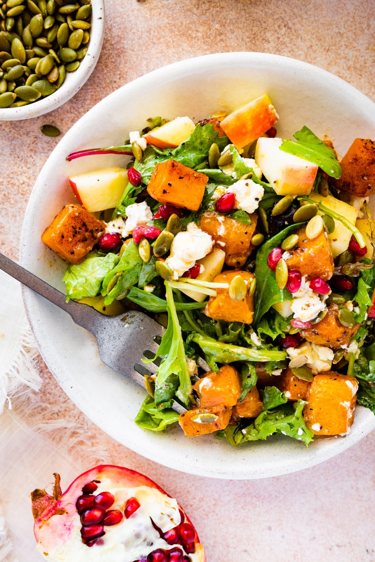 Thanksgiving salad in a small bowl with the ingredients that include butternut squash, apple, goat cheese, pomegranate, pumpkin seeds, and greens.