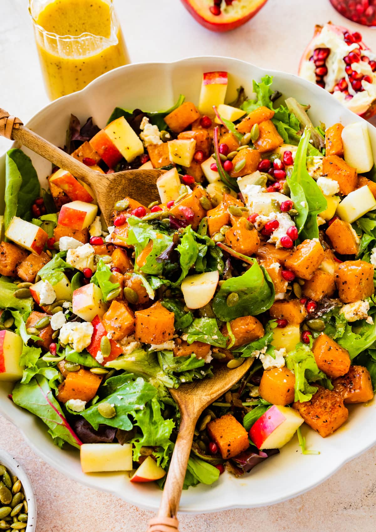 Thanksgiving salad in a large bowl with two wooden serving spoons. Ingredients include butternut squash, apple, goat cheese, pomegranate, pumpkin seeds, and greens.