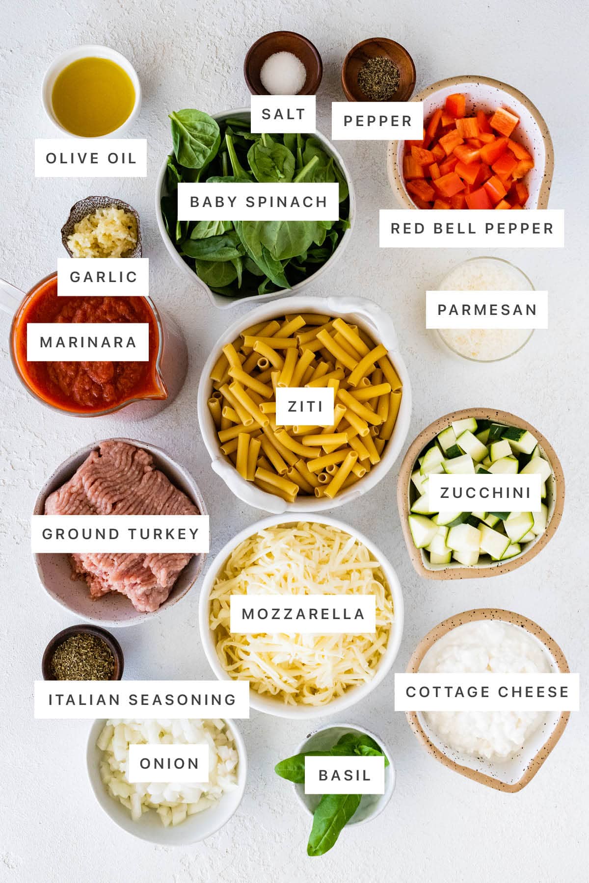 Ingredients measured out to make Easy Baked Ziti: olive oil, salt, pepper, baby spinach, red bell pepper, garlic, marinara, ziti, parmesan, ground turkey, mozzarella, zucchini, Italian seasoning, onion, basil and cottage cheese.