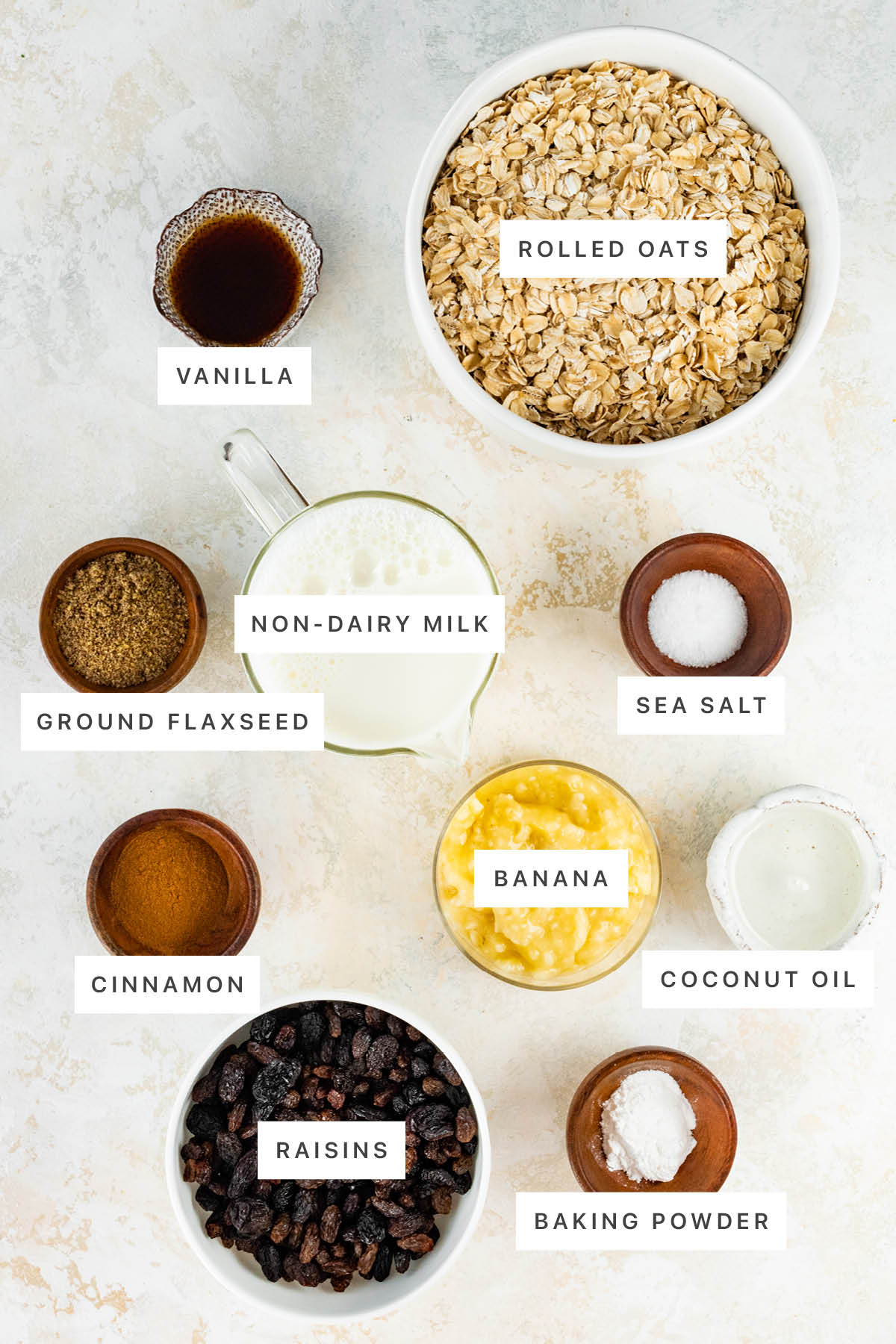 Ingredients measured out to make Cinnamon Raisin Baked Oatmeal: vanilla, rolled oats, non-dairy milk, ground flaxseed, sea salt, cinnamon, banana, coconut oil, raisins and baking powder.