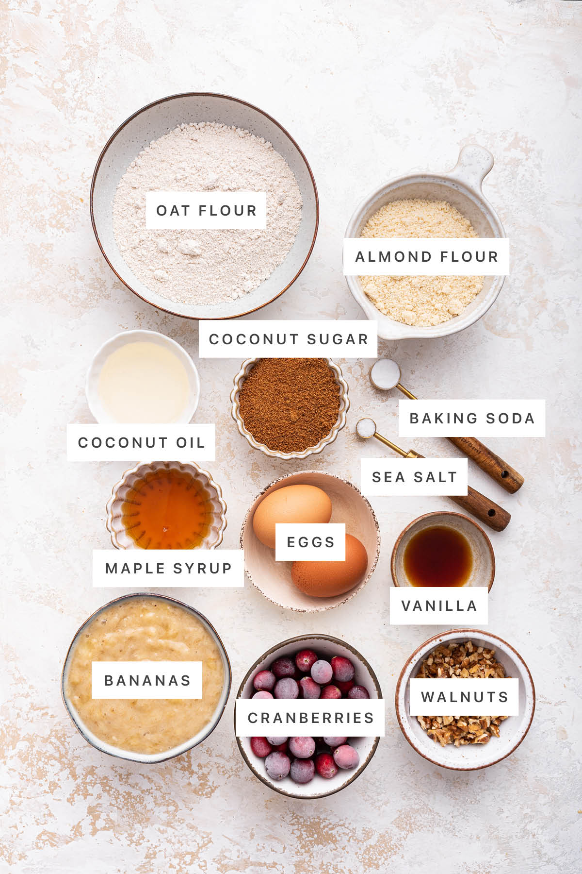 Ingredients measured out to make Cranberry Banana Bread: oat flour, almond flour, coconut sugar, coconut oil, baking soda, sea salt, maple syrup, eggs, vanilla, bananas, cranberries and walnuts.