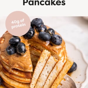 Stack of Protein Pancakes with a bite taken out of it with a fork. Stack of pancakes are topped with peanut butter, maple syrup and blueberries.