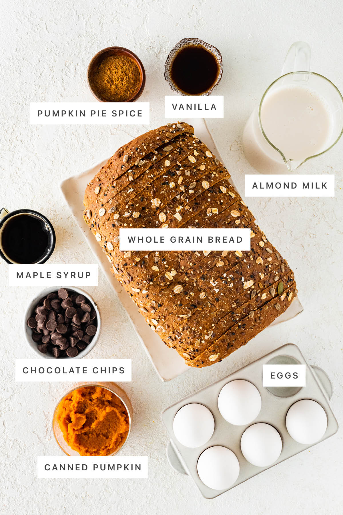 Ingredients measured out to make Pumpkin French Toast Casserole: pumpkin pie spice, vanilla, almond milk, whole grain Bread, maple syrup, chocolate chips, canned pumpkin and eggs.