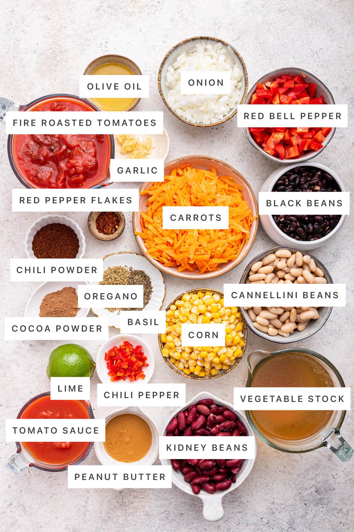 Ingredients measured out to make Easy Vegetarian Chili: fire roasted tomatoes, olive oil, onion, red bell pepper, garlic, red pepper flakes, carrots, black beans, chili powder, oregano, cocoa powder, basil, corn, cannellini beans, lime, chili pepper, tomato sauce, peanut butter, kidney beans and vegetable stock.