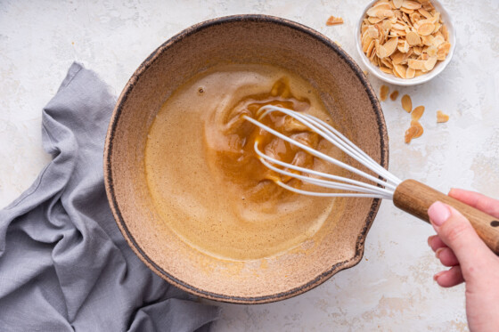 A woman's hand uses a whisk to whisk the wet ingredients for the almond cake in a large bowl.