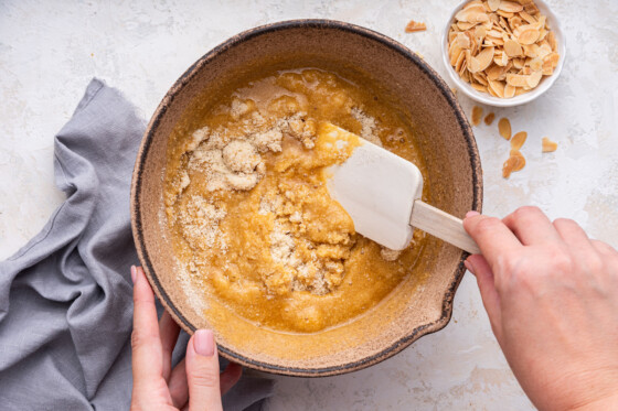 A woman's hand uses a silicone spatula to fold the wet and dry ingredients used for an almond cake in a large bowl.