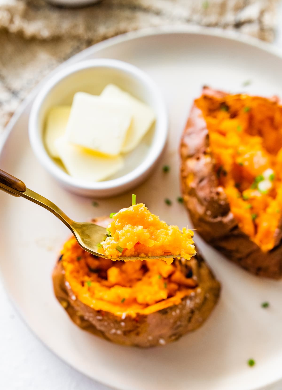 A fork holding a bite size piece of sweet potato above a plate with two baked sweet potatoes on it.
