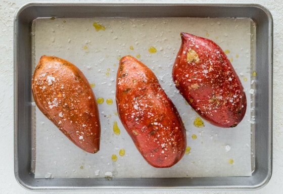 Three large sweet potatoes covered in oil and salt on a baking tray.