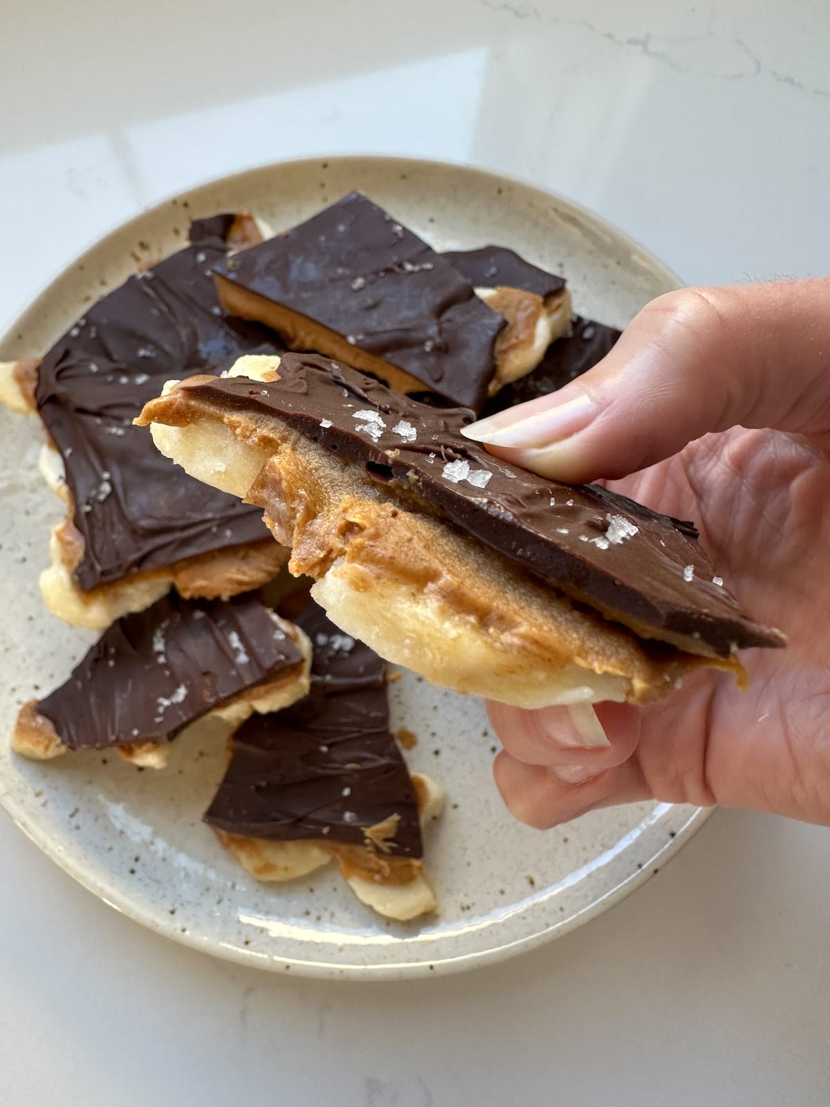 A woman's hand holding a piece of banana bark with layers of peanut butter and chocolate.