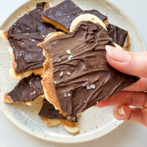 A woman's hand holding a piece of banana bark with layers of peanut butter and chocolate with flaky sea salt.
