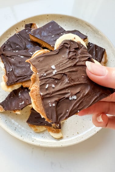 A woman's hand holding a piece of banana bark with layers of peanut butter and chocolate with flaky sea salt.