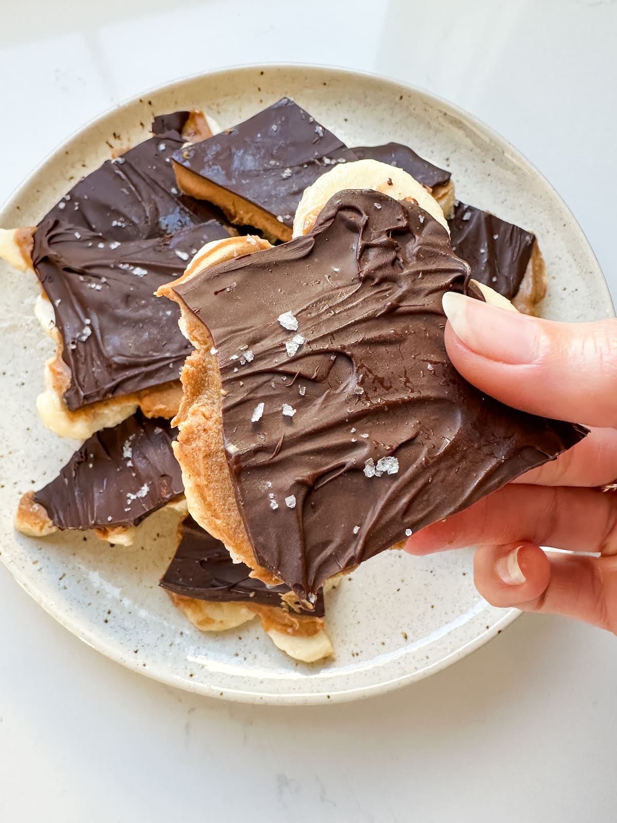 A woman's hand holding a piece of banana bark with layers of peanut butter and chocolate with flaky sea salt.