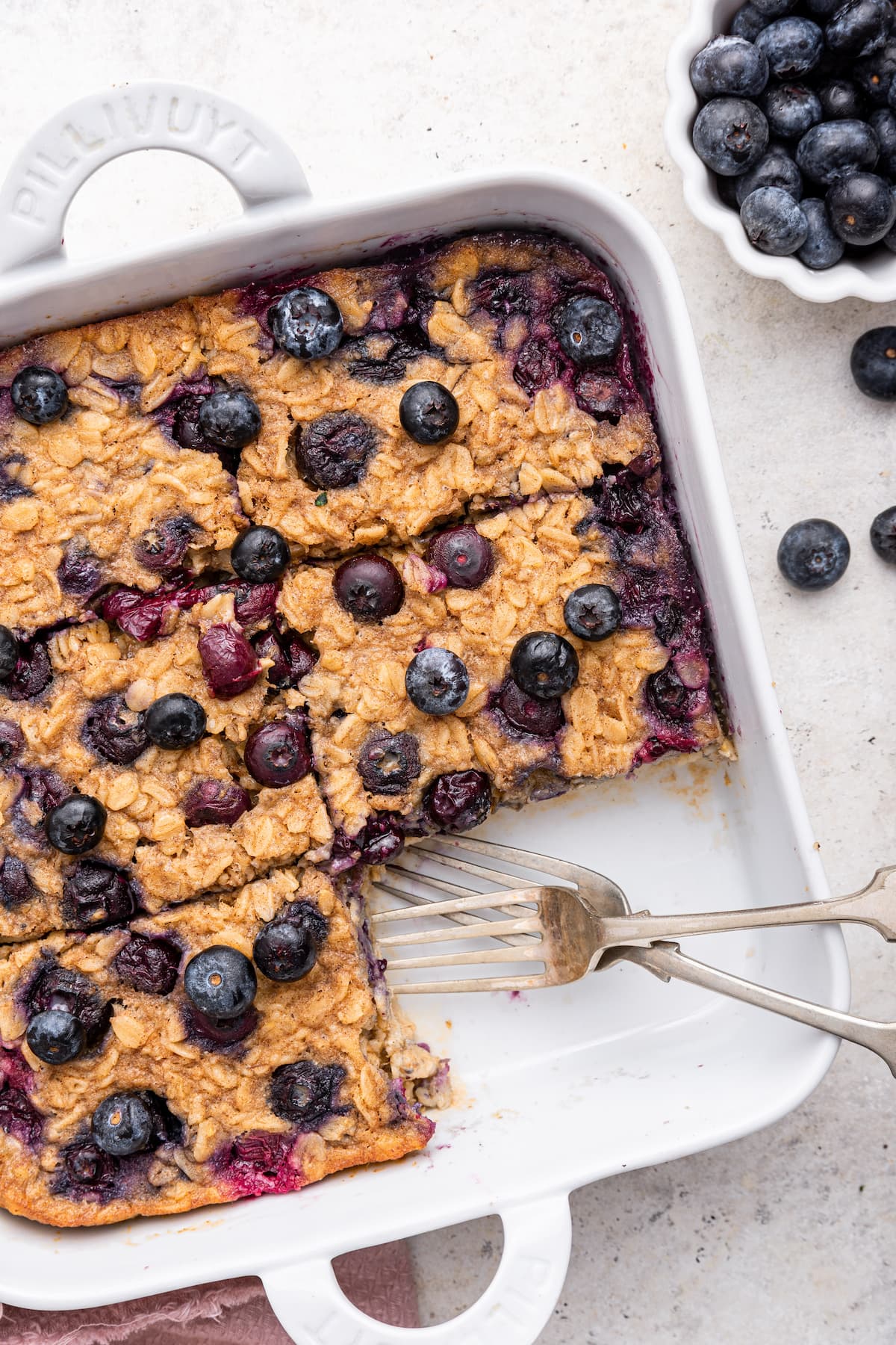 Blueberry baked oatmeal in a square baking dish with a piece taken from the dish. There are two metal forks in place of the missing piece.