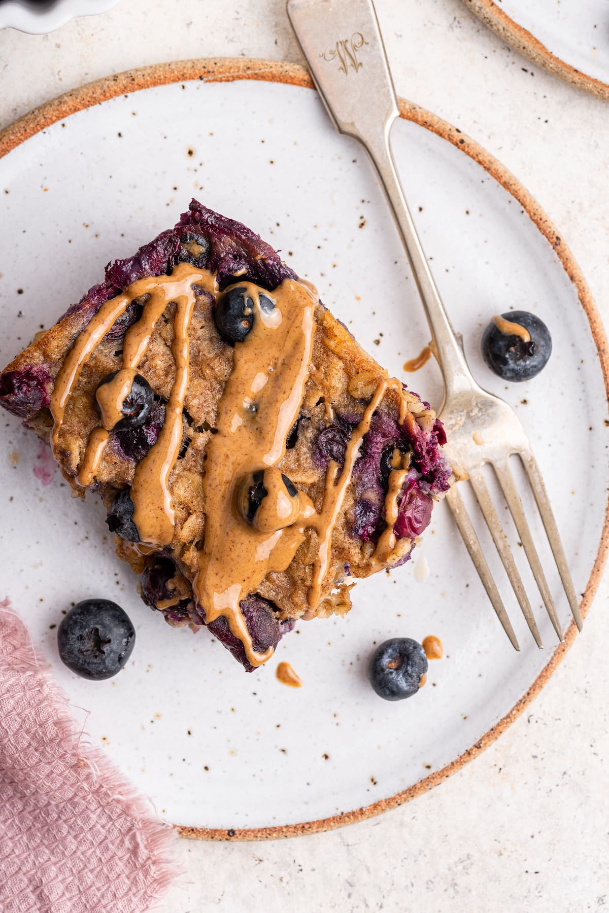 A serving of blueberry baked oatmeal on a small plate with a metal fork. The square piece of baked oatmeal has a drizzle of nut butter on top.