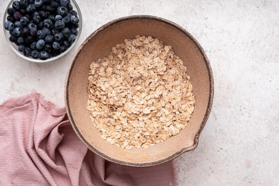 Rolled oats in a large brown mixing bowl near a small bowl of blueberries.