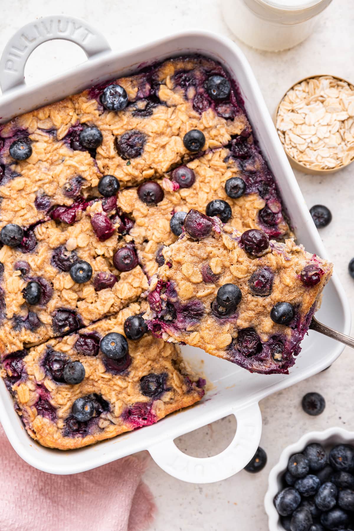 A serving of blueberry baked oatmeal being lifted away from a square baking dish with the rest of the baked oatmeal by a fork.