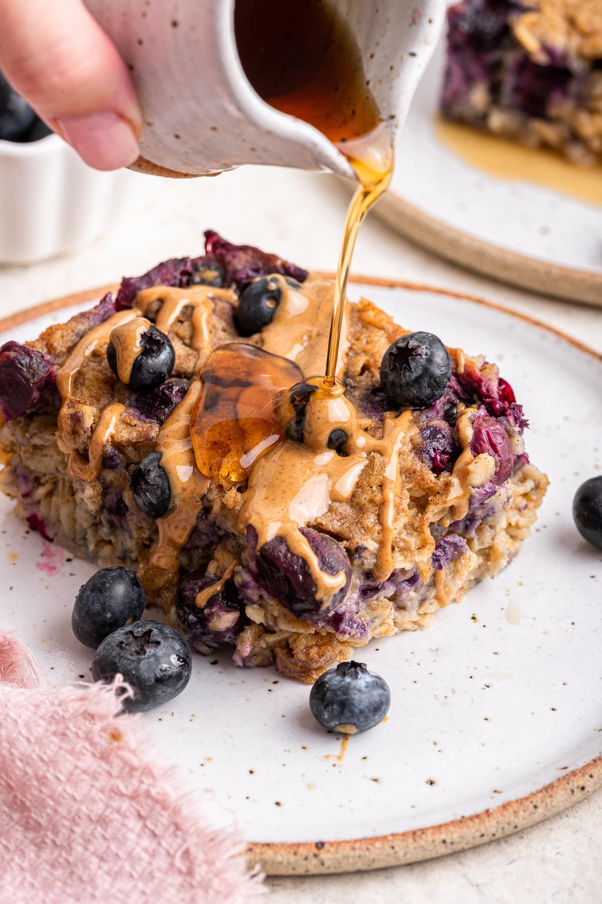 Maple syrup is being poured over a serving of blueberry baked oatmeal that also has a drizzle of nut butter.