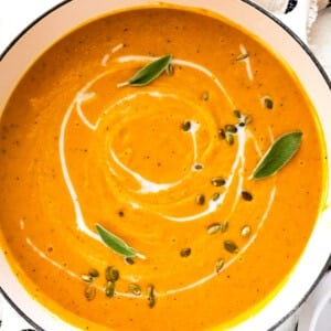 Butternut squash soup in a large pot garnished with coconut milk, pumpkin seeds, and fresh sage.