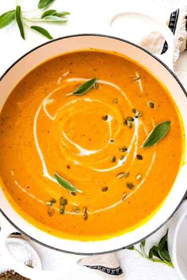 Butternut squash soup in a large pot garnished with coconut milk, pumpkin seeds, and fresh sage.