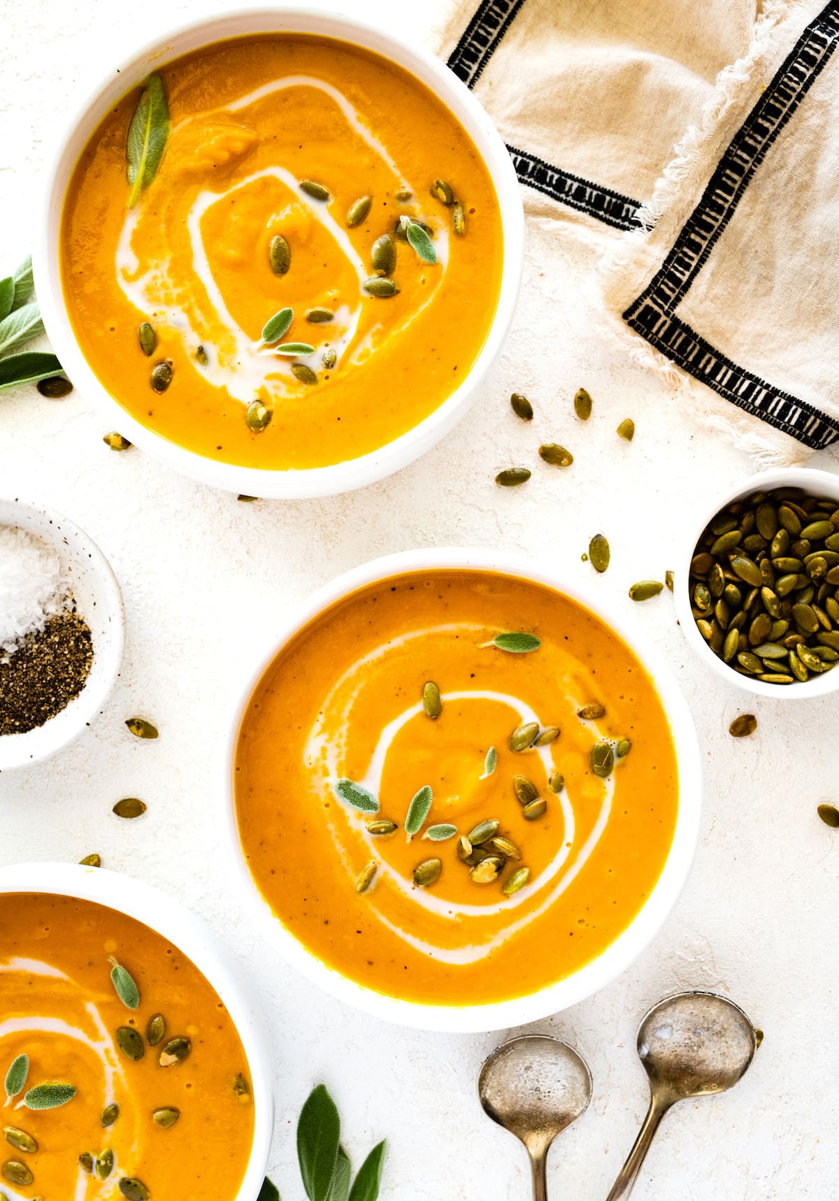 Butternut squash soup in three bowls garnished with coconut milk, pumpkin seeds, and fresh sage.